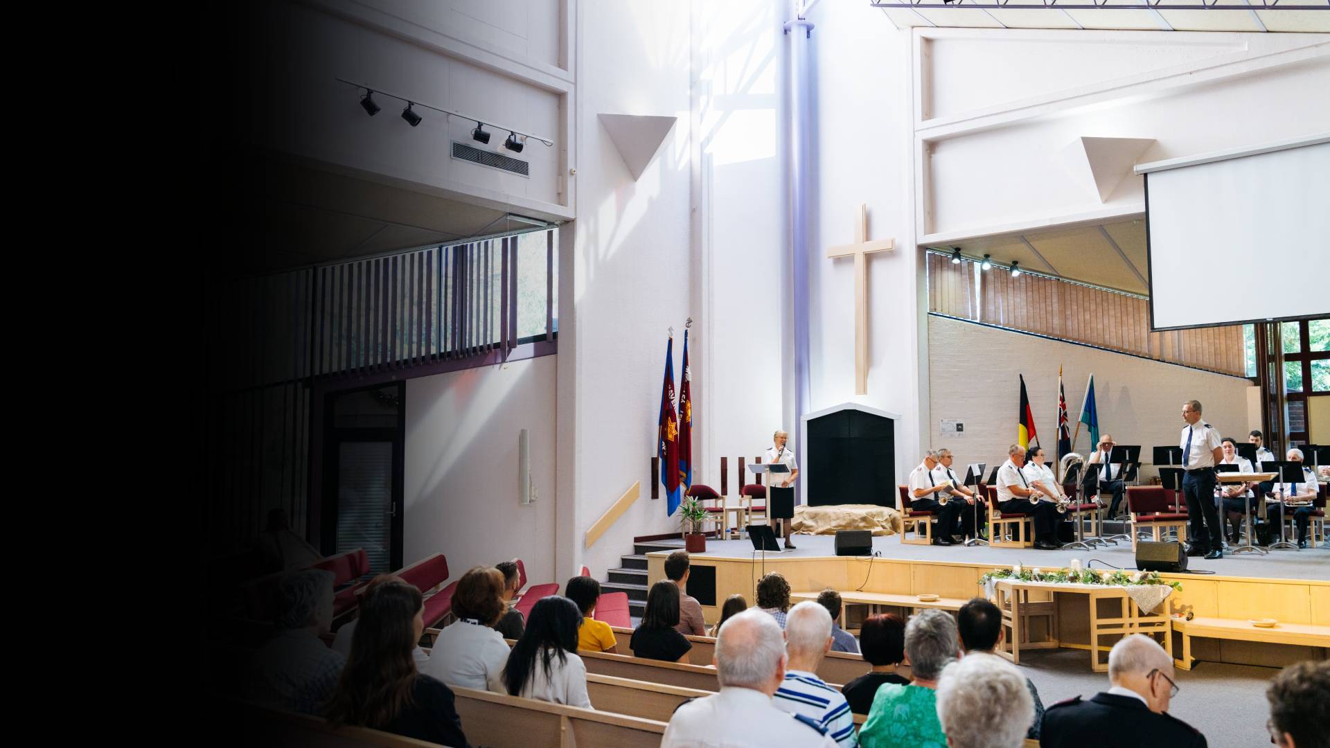 A wide view a brightly lit church service. An officers speaks from the stage to the congregation in our foreground.