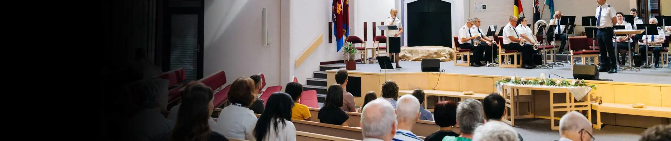 A wide view a brightly lit church service. An officers speaks from the stage to the congregation in our foreground.