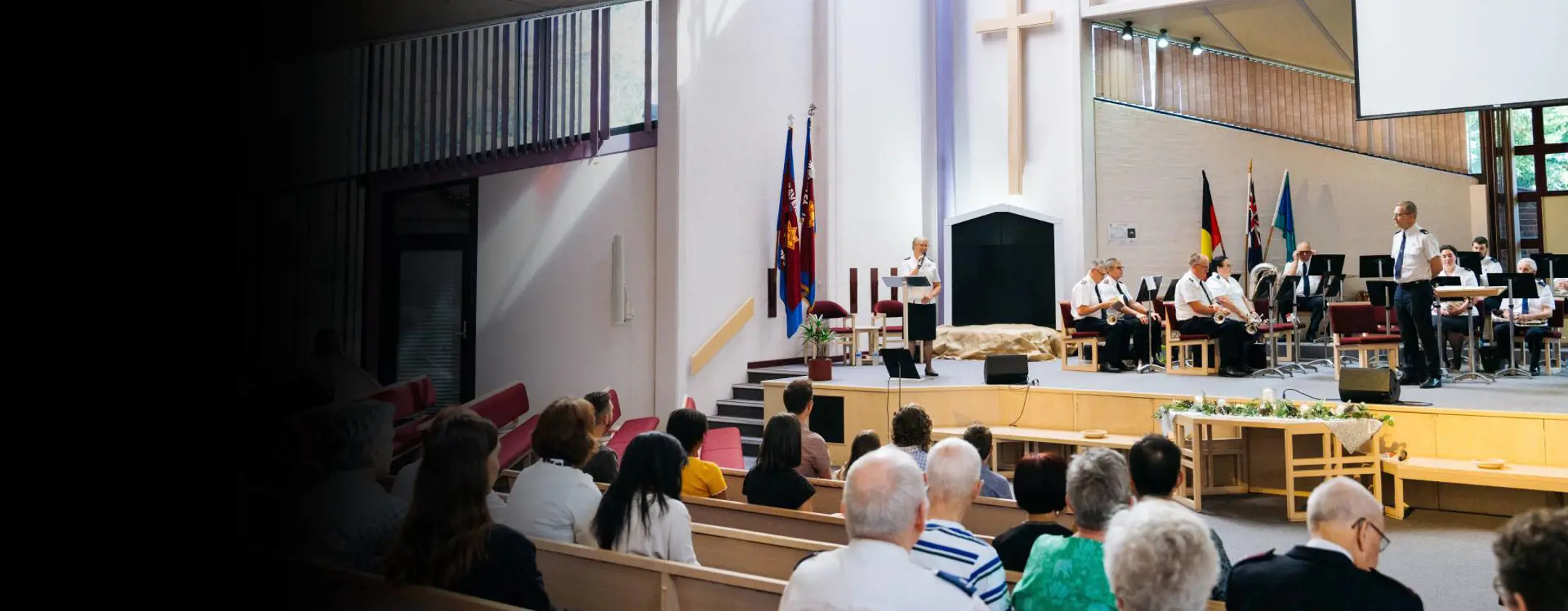 A wide view a brightly lit church service. An officers speaks from the stage to the congregation in our foreground.