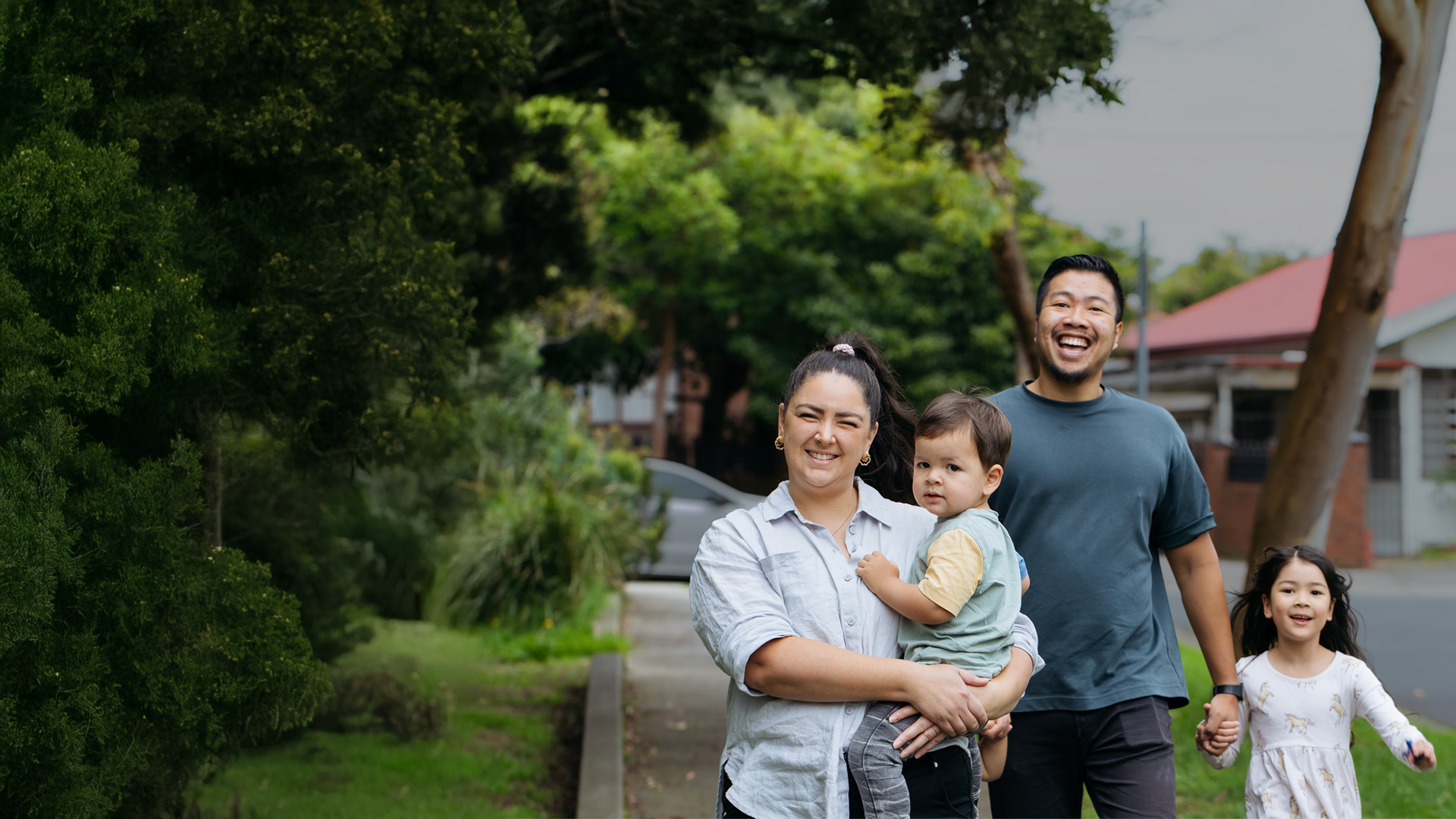 Smiling young family of four enjoying a walk along a leafy suburban path — mum cradling the toddler while dad gently holds their daughter’s hand.
