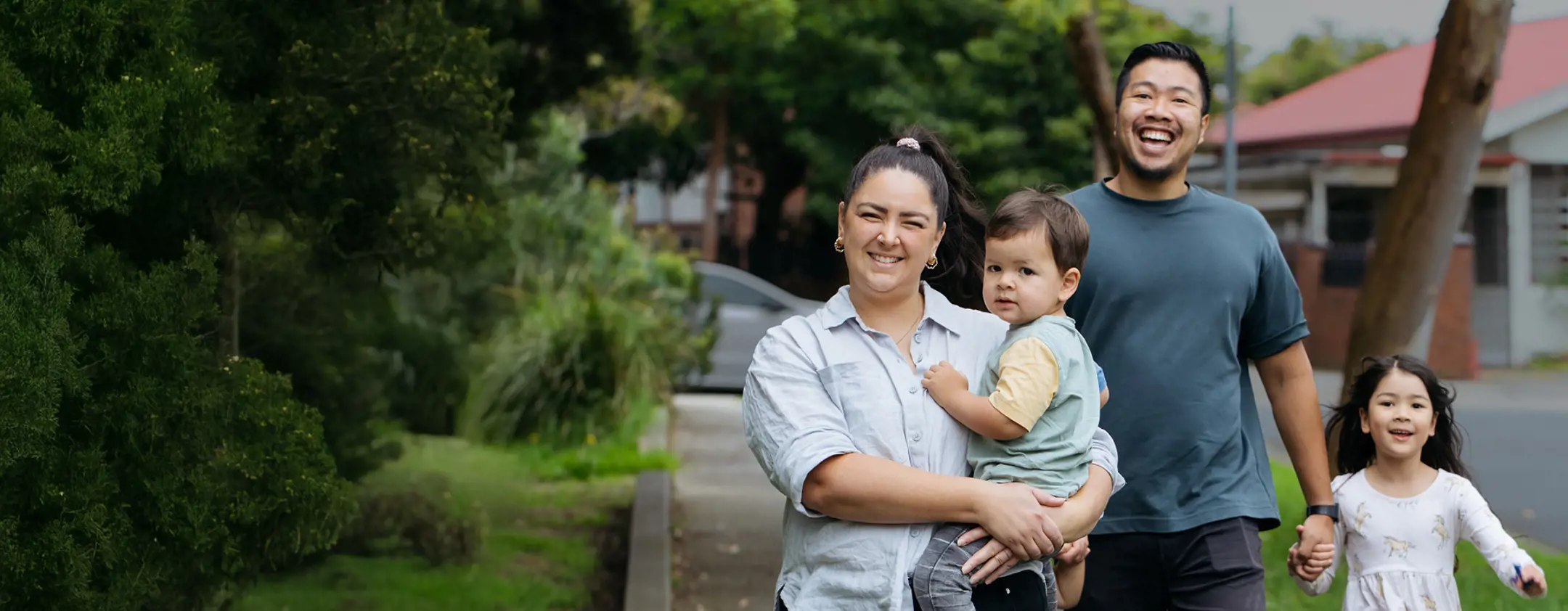 Smiling young family of four enjoying a walk along a leafy suburban path — mum cradling the toddler while dad gently holds their daughter’s hand.