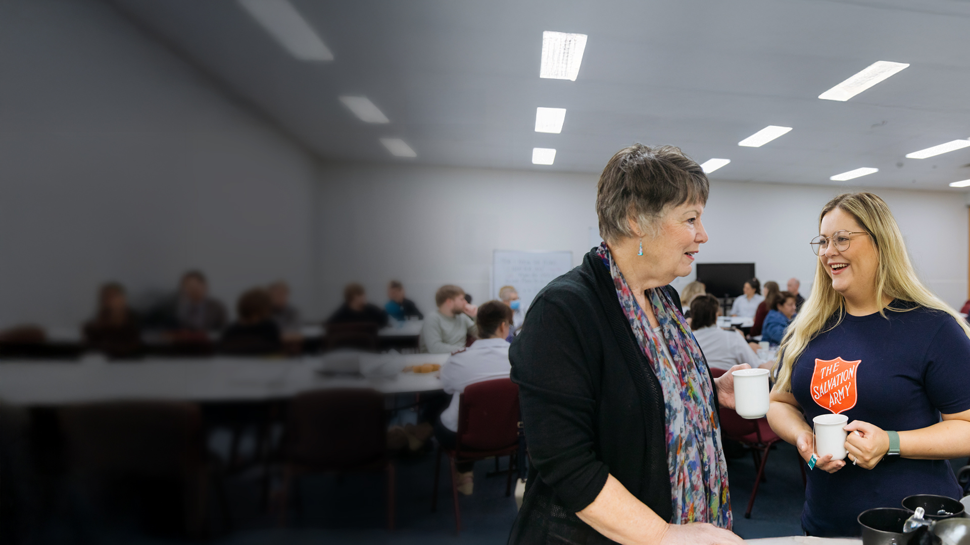 Two women share a warm cup of tea and friendly conversation in a lively room filled with people socialising — one wearing a Salvation Army t-shirt.