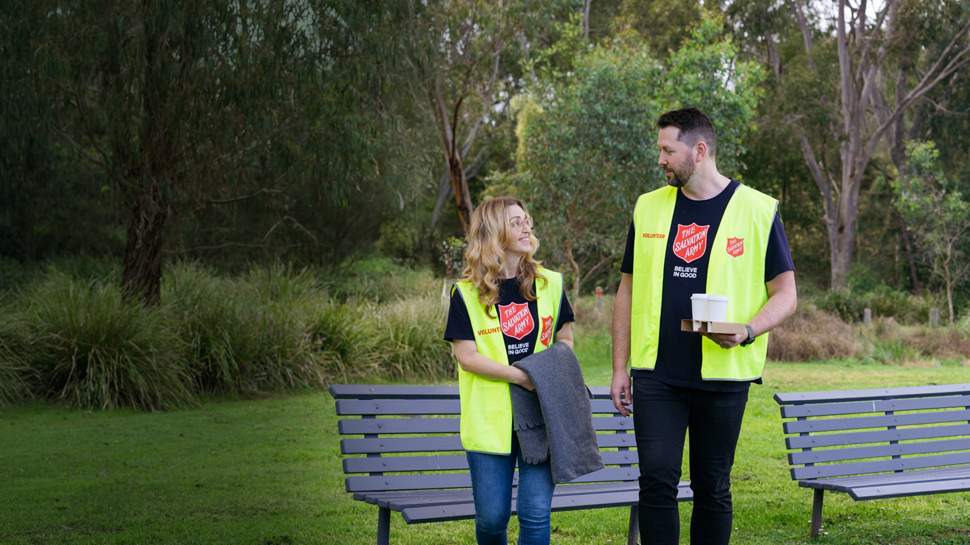 Two Salvation Army volunteers sharing a friendly conversation in a park, ready with warm coffee, blankets, and open hearts to support those doing it tough.