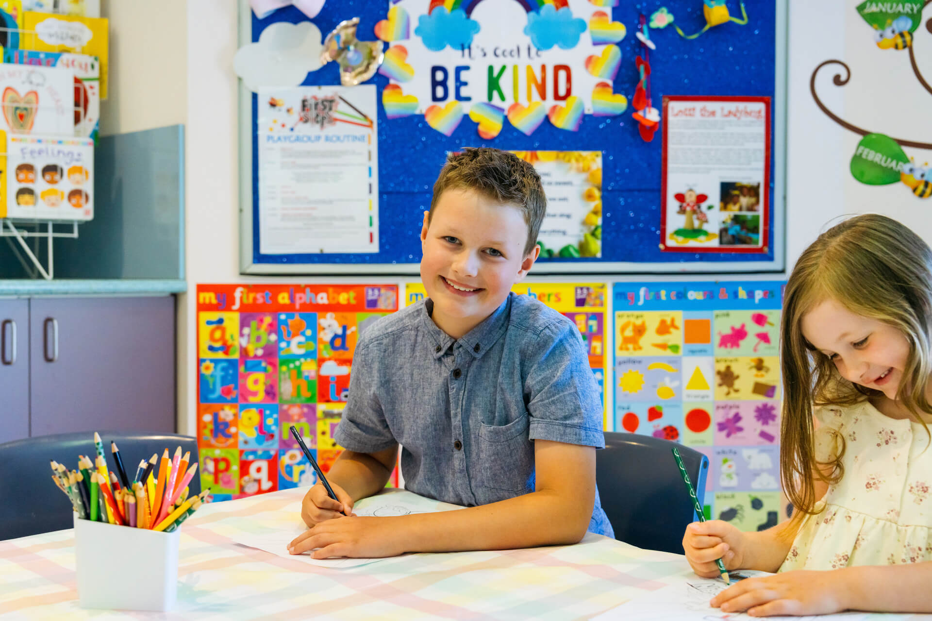 Two children sitting at a table smiling while drawing on paper.