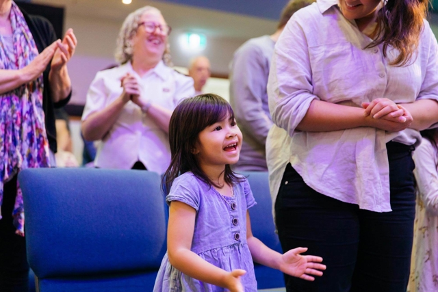 A young girl sings and claps enthusiastically during a church service with her family.